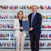 A formal meeting captured in a photograph with two individuals holding a document in front of a wall filled with flags.