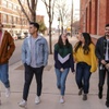 A group of five young adults walking and talking on a city sidewalk next to a brick building.