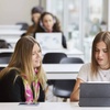 Two women collaborating on a work project with laptops at a white desk in an office environment.