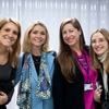 Four women smiling at the camera in an office-like environment with a white curtain in the background.
