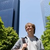A young man stands smiling in front of modern skyscrapers on a sunny day.