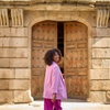 A woman in a pink suit smiles while walking past an old wooden door of a stone building.