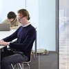 A young man is using a laptop in a modern office environment with other people working in the background.