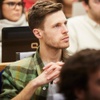 A young man with a green plaid shirt is listening attentively in a classroom setting.