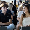 A young man and woman smiling and talking, sitting in a room filled with people.