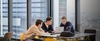 Three young men are collaborating on a laptop at a modern workspace with a city view.