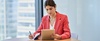 A woman in a pink blazer is working on an iPad while taking notes at a table.