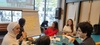 A group of diverse people engaged in a discussion around a table with notes and a whiteboard in a bright room.