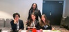 A group of four women in professional attire are seated at a table in a conference setting.