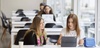 Two women collaborating on a work project with laptops at a white desk in an office environment.