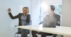 A woman writing on a whiteboard while a man watches, both seated in a modern office setting.