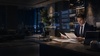 A man in a suit is reading documents at a modern office desk in a dimly lit environment.