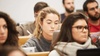 A focused young woman using a laptop in a classroom full of students.
