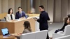 A man is speaking at a podium in a modern conference room with several attendees and a panel of two men and a woman.