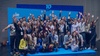A large group of cheerful people celebrating together in front of a blue backdrop.