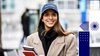 A smiling woman wearing a university cap and a beige coat holds books, standing outdoors.