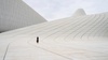 A solitary figure walks on a smooth, curving surface of a modern architectural structure.
