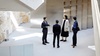 Four business professionals standing and conversing in a modern architectural setting with large columns and natural light.