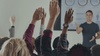 A group of people raising their hands in a classroom setting during a presentation.