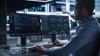 A man working at a computer desk with dual monitors displaying data analytics and charts.