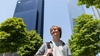 A young man stands smiling in front of modern skyscrapers on a sunny day.