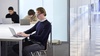 A young man is using a laptop in a modern office environment with other people working in the background.