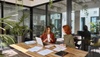 Two women discussing documents at a wooden table in a modern office with natural light and plants.
