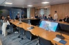 A professional meeting is in session in a conference room with multiple attendees and a presentation on a screen.