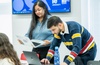 Two students are engaged in a discussion while working on their laptops in a classroom.