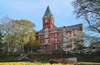 A historic brick building with a tower, surrounded by trees and flowering plants in a park-like setting.