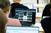 A group of people in a classroom setting, focusing on a laptop screen displaying various software applications.