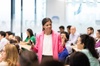 A woman in a pink blazer is speaking to a group of seated people in a bright modern office.