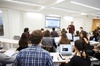 A lecturer explaining to students in a classroom equipped with laptops and a projector screen.