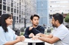 Three people having a conversation at a table outdoors, with modern buildings in the background.