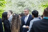 A man in a suit with a tie is smiling while holding a coffee cup, interacting with other people at an outdoor gathering.