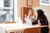 Two women are engaged in a conversation while working on their laptops at a table.