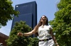 A smiling woman walking among green trees with a modern building in the background.