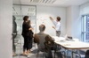 A team meeting in a modern office space with a person presenting on a whiteboard.
