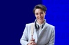 A smiling young woman in a stylish gray coat with headphones around her neck, set against a vibrant blue background.