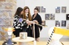 Three women examining a piece of fabric in an art gallery with various artworks displayed in the background.