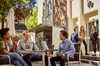 Three students are having a discussion at an outdoor seating area of a campus, with other individuals passing by.