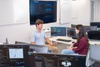 Two students are discussing at a table with multiple computer screens displaying data analysis.