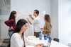 A group of four young people collaborate in a modern workspace, with one drawing on a whiteboard while others engage in discussion.