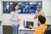 A group of three people having a discussion in an office setting with digital screens displaying data in the background.