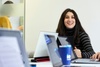 A smiling young woman sitting at a table with laptops and a coffee cup around her.