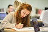 A student is focused on writing while studying in a busy environment.
