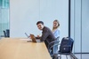 Two young adults are sitting at a table in a modern workspace, smiling and engaging with their laptops.