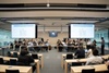 A group of people seated in a modern conference room with presentations displayed on screens.