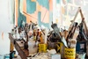 A cluttered artist's table filled with paint brushes, open paint cans, and various painting supplies in front of a colorful abstract painting.