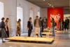 People viewing an art exhibition in a gallery with red and white walls and modern artwork installations.
